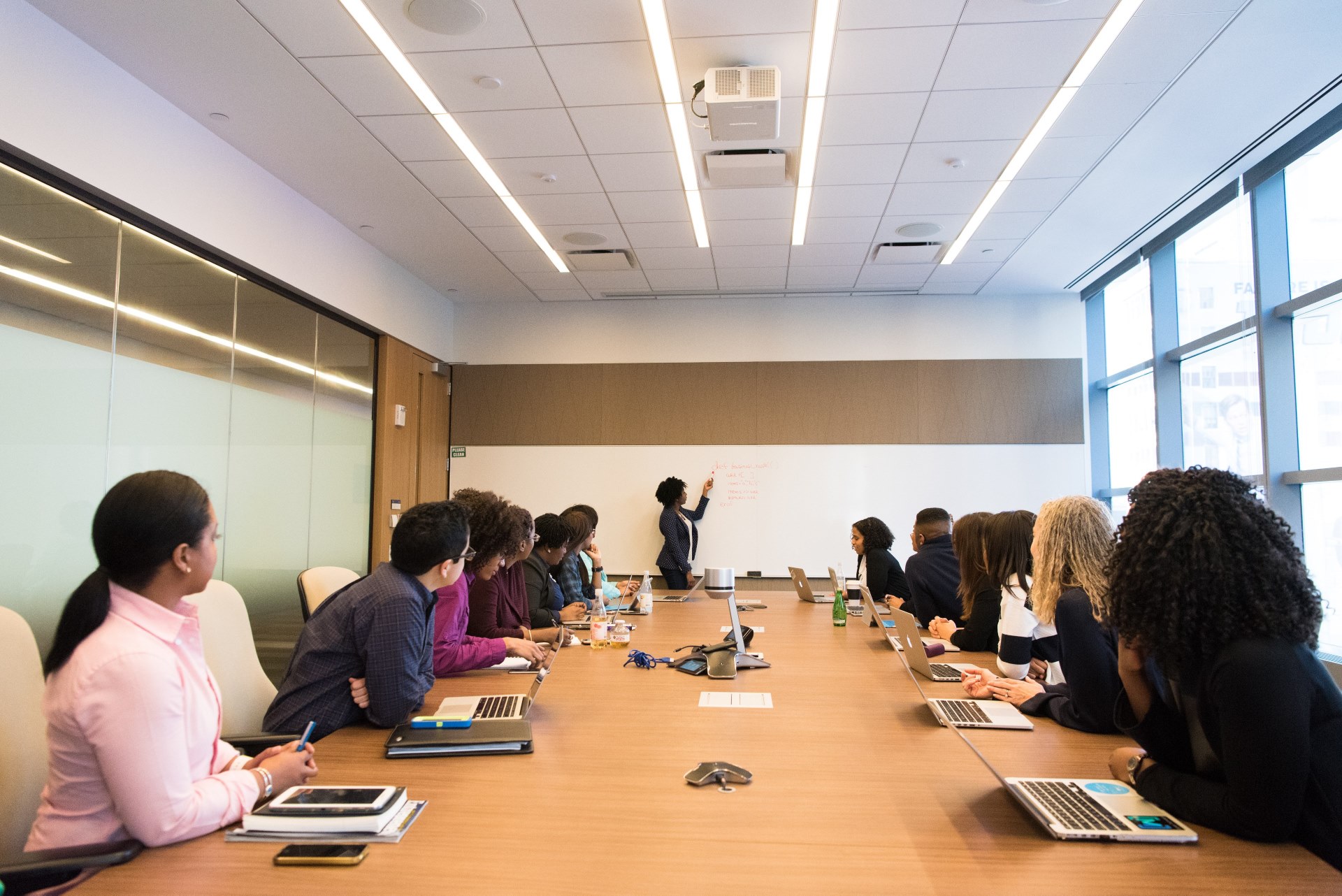 Photo of a group of people in a meeting room with one person at the whiteboard writing something
