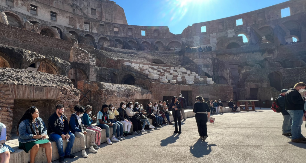 group of students sitting inside what looks like the Rome Colosseum with a bright sun shining down from the right top corner.