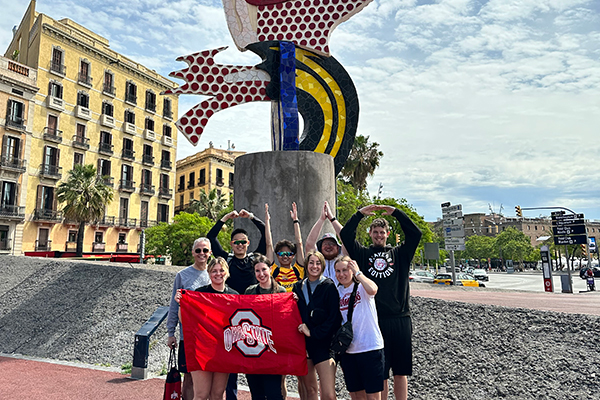A group of smiling students standing in front of a very colorful sculpture, some of the students are holding an Ohio State Flag, with The Ohio State University's athletic logo on it.