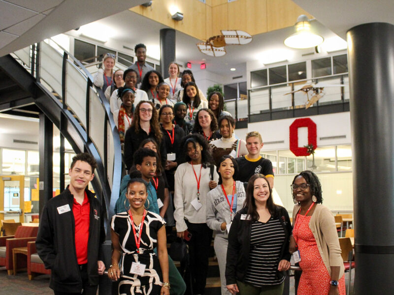 ROOT Participants and program leaders stand on stairs facing camera, smiling. A large scarlet block O is in the background.