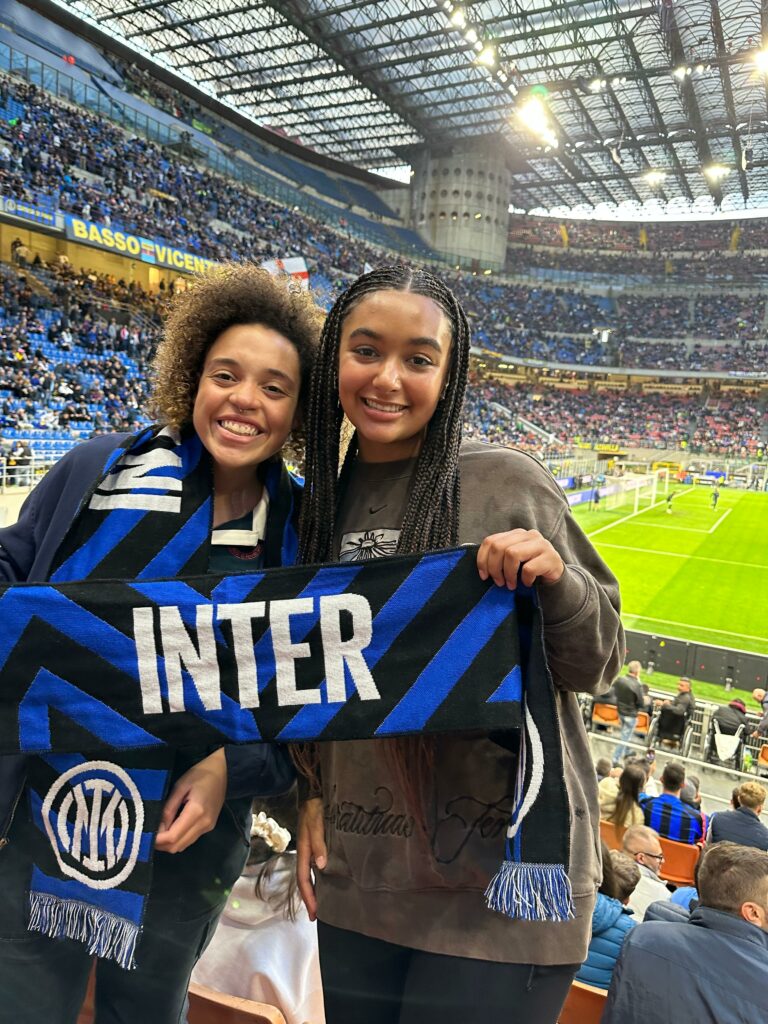 Two women stand in front of a stadium filled with people